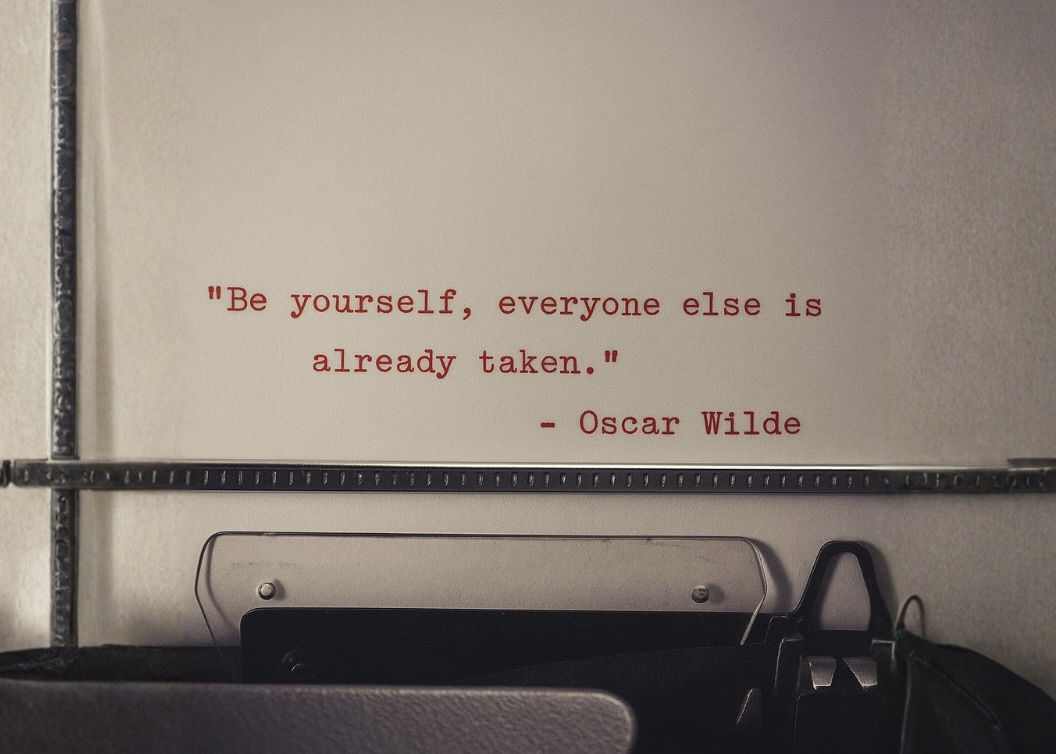 Close-up of a typewriter typing the quote “Be yourself, everyone else is already taken.” — Oscar Wilde, on white paper with a reddish tint.