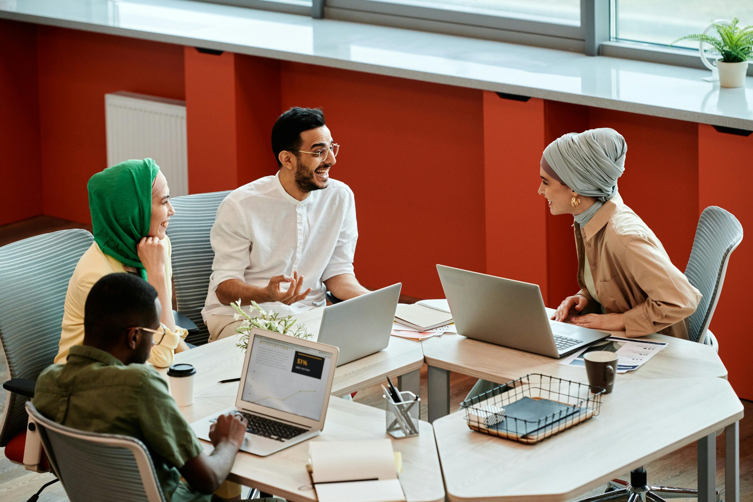 Four people sit around a table with laptops, engaged in a lively discussion. The atmosphere is collaborative and friendly, set in a modern office.