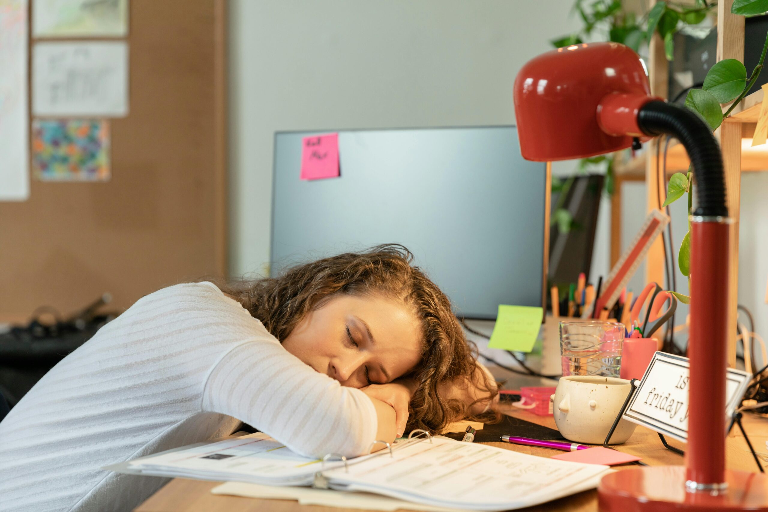 A woman with curly hair is asleep at her desk, resting her head on her arms beside open notebooks, a computer monitor, and a red desk lamp.